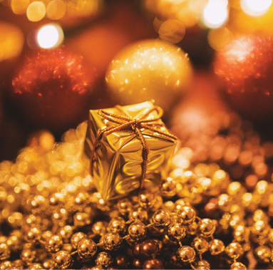 small gift box wrapped in gold wrapping paper with gold string, sitting atop gold beads, blurred background of red and gold ornaments